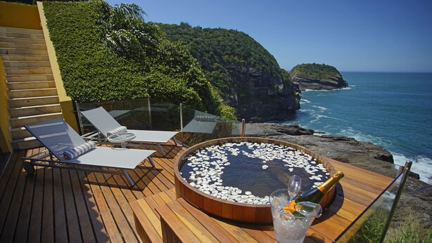 Terraza de madera con sillones de descanso y jacuzzi, rodeada por una barandilla de cristal, con vistas a un acantilado costero y al océano.