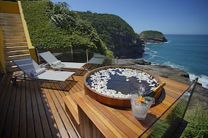 A wooden deck with lounge chairs and a hot tub surrounded by a glass railing, overlooking a coastal cliff and ocean.