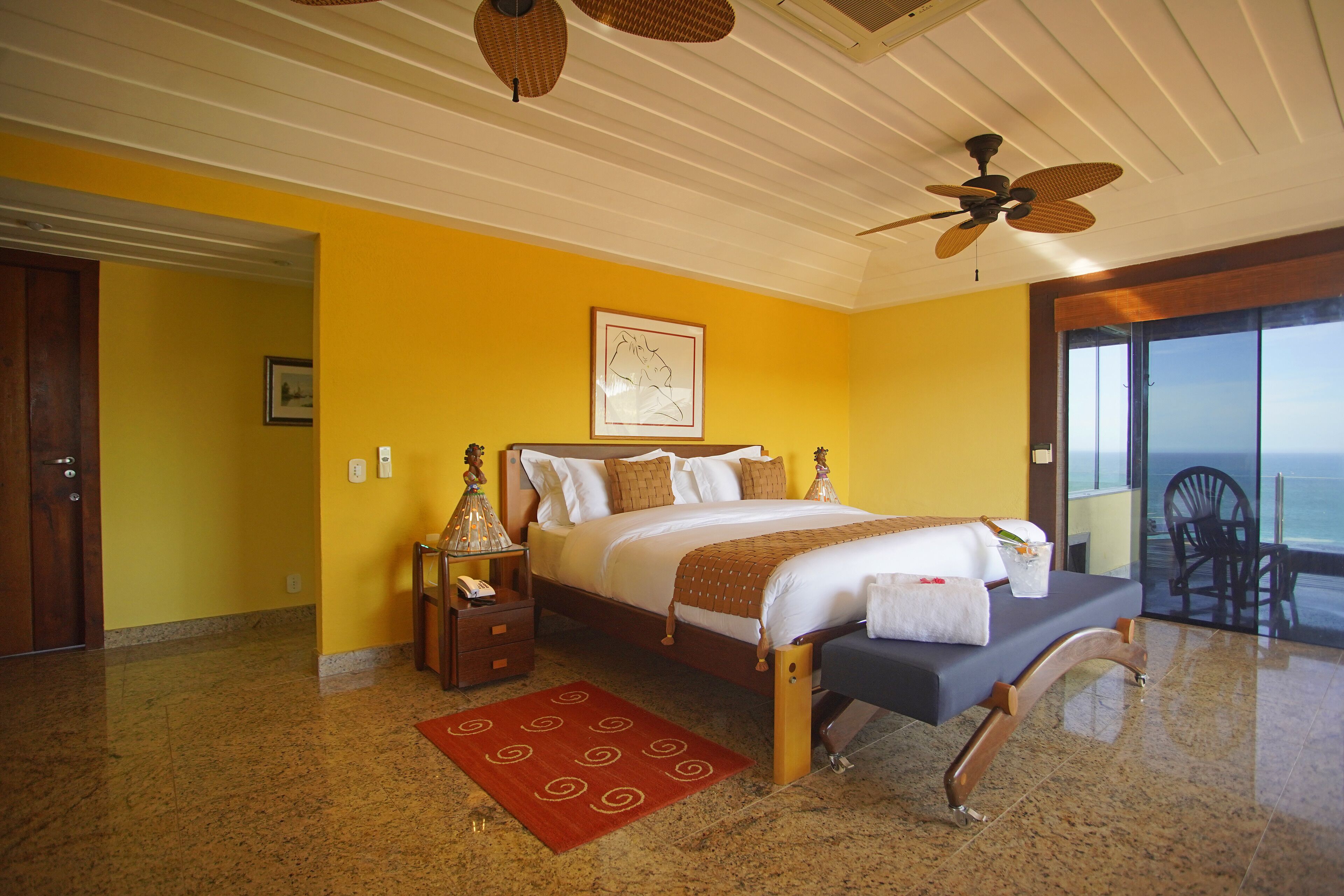 A bedroom with a bed, a nightstand, a red rug, a ceiling fan, and a large window with a view.