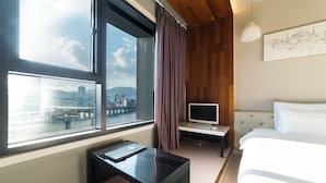 Une chambre d’hôtel moderne dotée d’une grande fenêtre, d’un lit, d’un bureau avec un ordinateur et offrant une vue sur le paysage urbain.