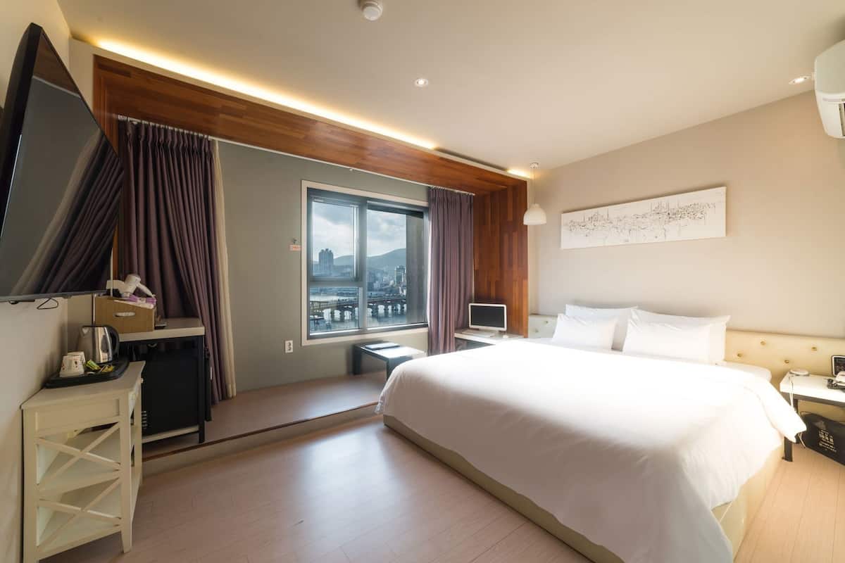 Habitación de hotel con cama, escritorio con computadora, televisor, ventana con vista a la ciudad y una obra de arte colgada en la pared.