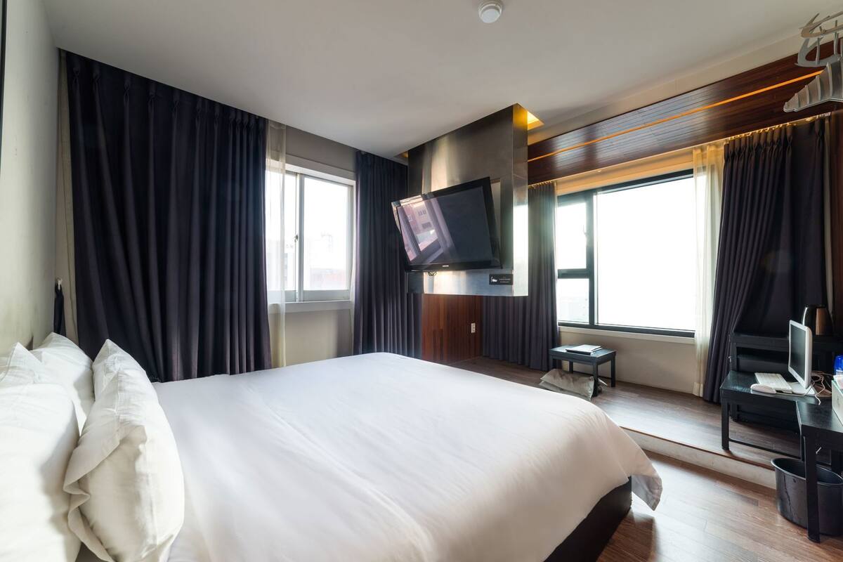 Una habitación de hotel con una cama grande, un televisor de pantalla plana, un escritorio con una computadora y una ventana con cortinas.