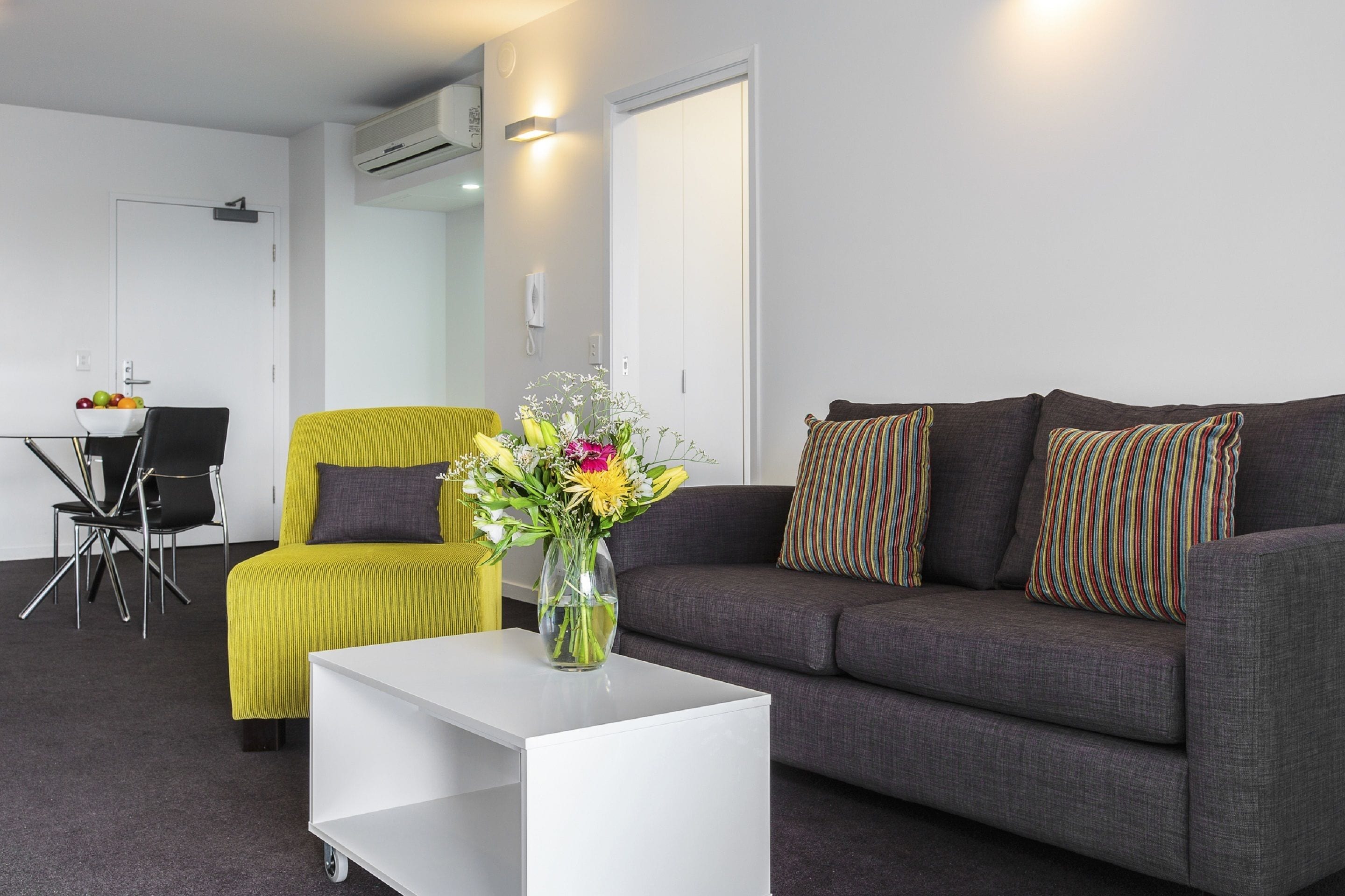 Een moderne woonkamer met een grijze bank, een gele fauteuil, een witte salontafel en een bloevas.