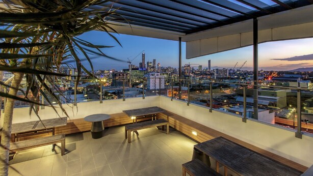 A rooftop terrace with a city view, wooden seating, and a glass railing. - Code Apartments (Bowen Hills)