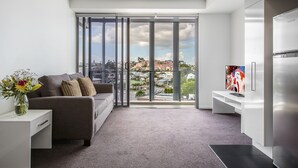 A modern living room with a grey sofa, a white side table with a vase, a large window with a city view, and a modern kitchen with a stainless steel refrigerator. - Code Apartments (Bowen Hills)