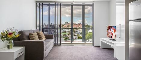 Una sala de estar moderna con un sofá gris, una mesita de noche blanca con un florero, un ventanal con vistas a la ciudad y una cocina moderna con un refrigerador de acero inoxidable.