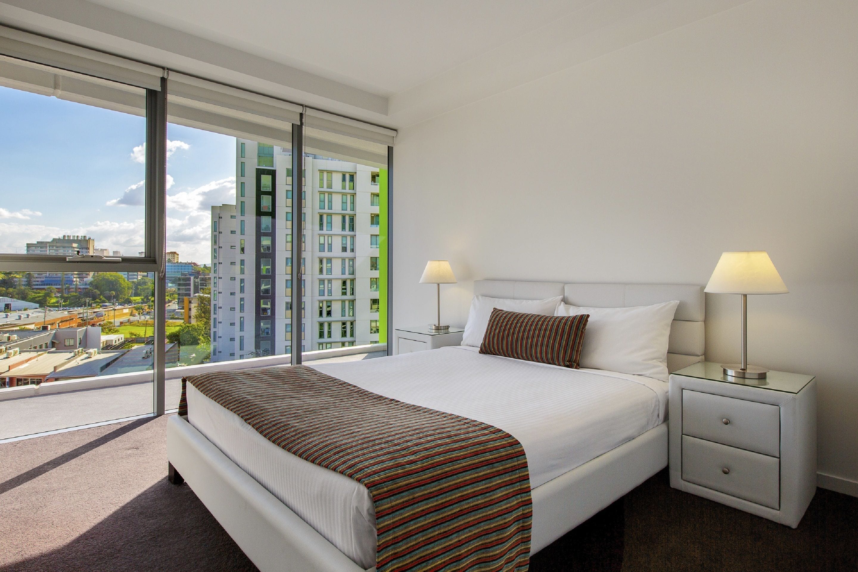 Ein modernes Hotelzimmer mit einem großen Bett, zwei Nachttischen inklusive Lampen, Blick auf eine Stadtkulisse durch raumhohe Fenster und einem Balkon.