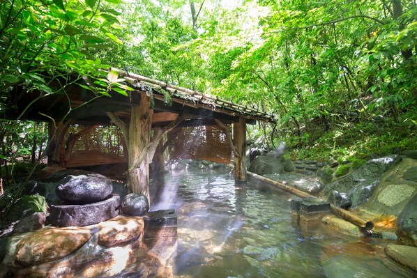 A wooden bridge over a clear stream with rocks and a wooden structure on the left.