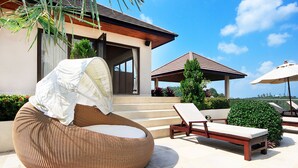 A modern outdoor seating area with a wicker lounge chair and a wooden bench, a covered patio, and a view of the house. - Baan Napoli Luxury Villa (Koh Samui)