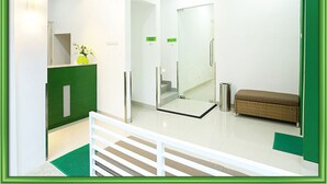 Zona de recepción con un mostrador verde, un banco blanco y una puerta de cristal que da a un pasillo.