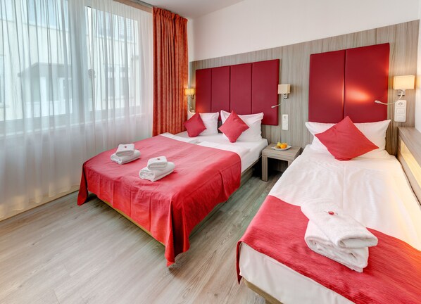 Habitación de hotel con dos camas, ropa de cama roja y blanca, una mesita de noche con lámpara y una ventana con cortinas traslúcidas.