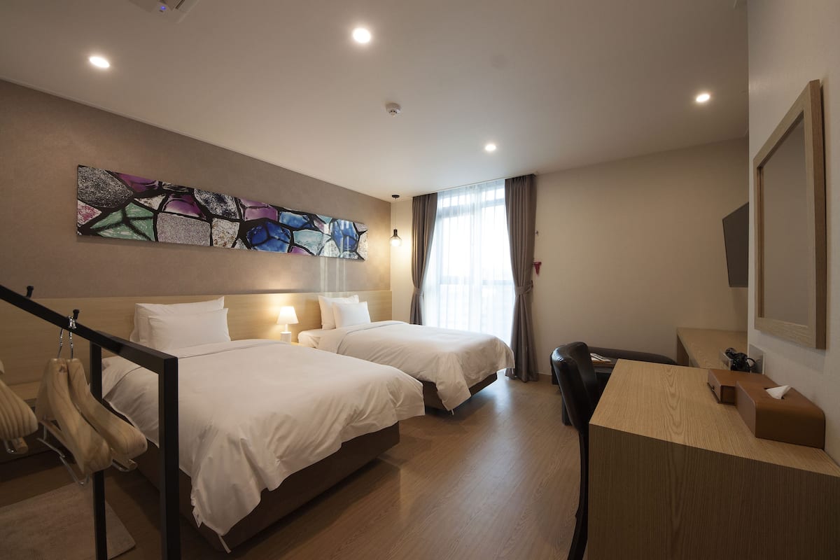 A hotel room with two beds, a desk, a chair, a large abstract painting, and a window with curtains.