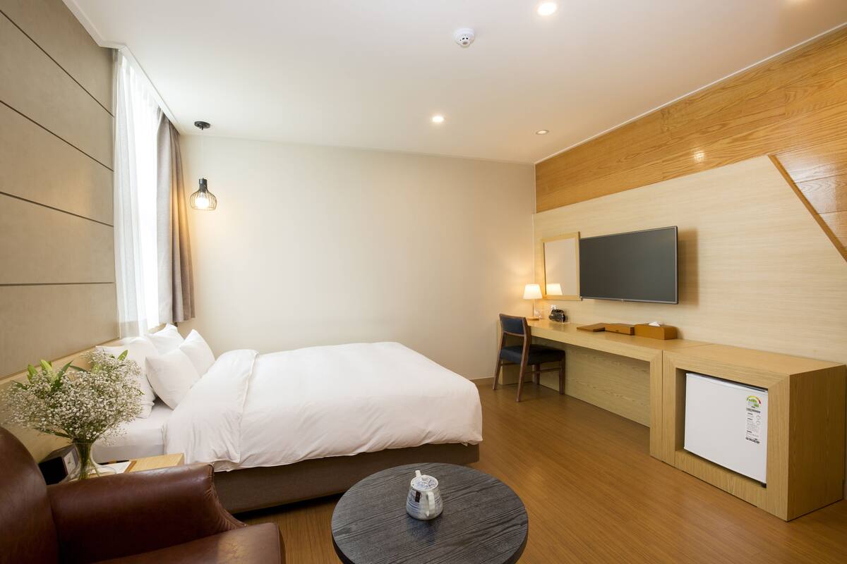 A hotel room with a bed, a desk, a chair, a coffee table, and a television mounted on the wall.