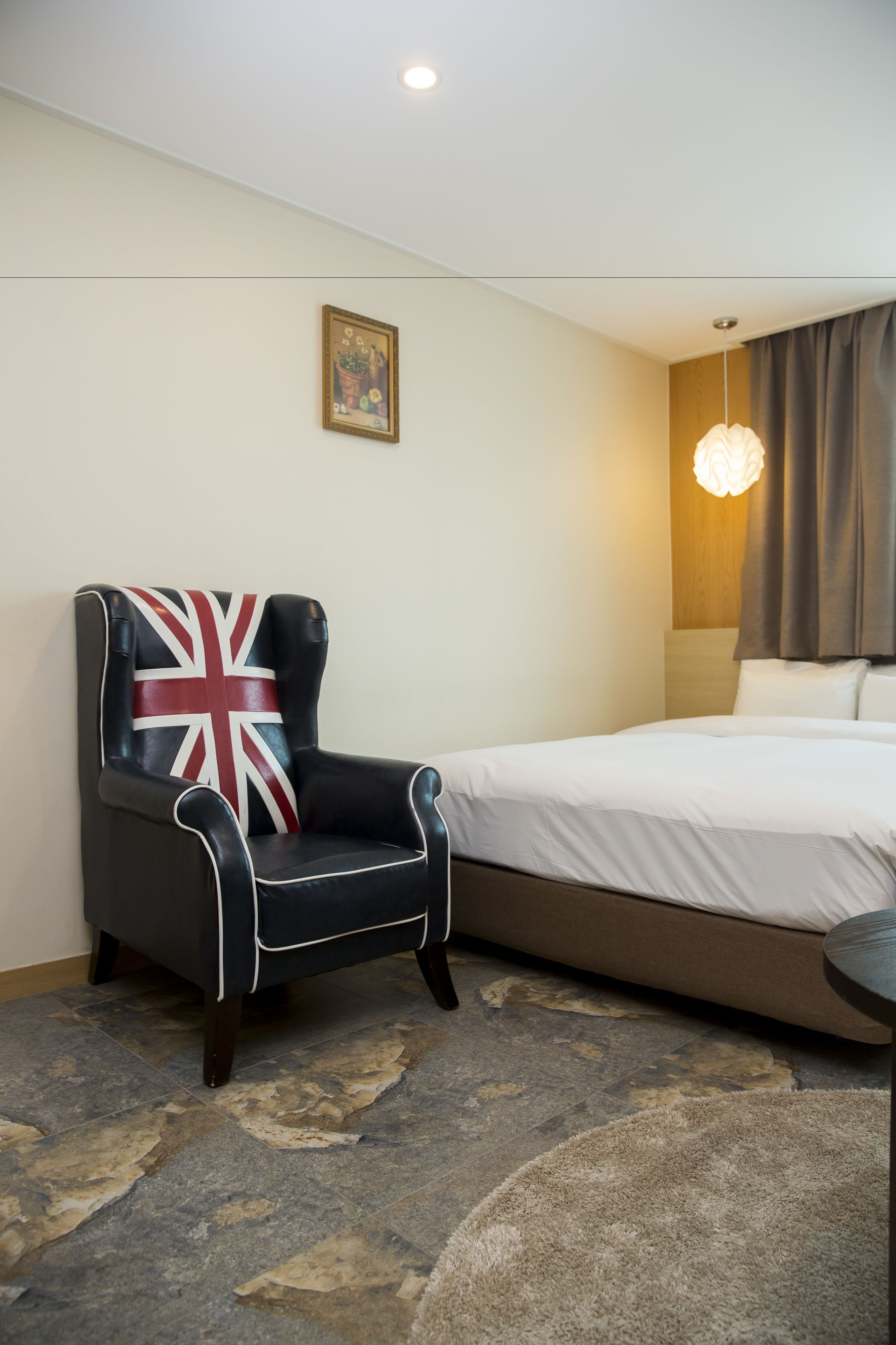 A hotel room with a bed, a chair with a Union Jack design, a painting on the wall, and a modern light fixture.