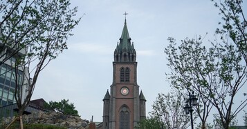 Una iglesia con una torre de reloj, rodeada de árboles y un muro de piedra.