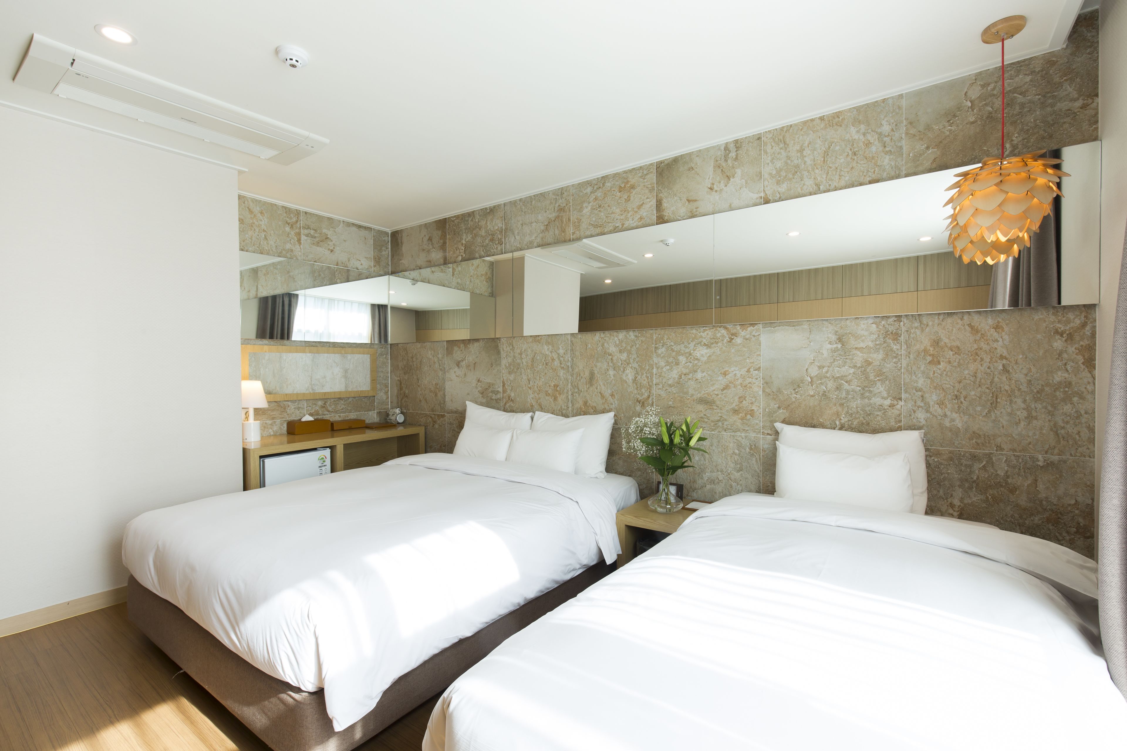 A hotel room with two beds, a bedside table with a lamp, a mirror, and a hanging light fixture.