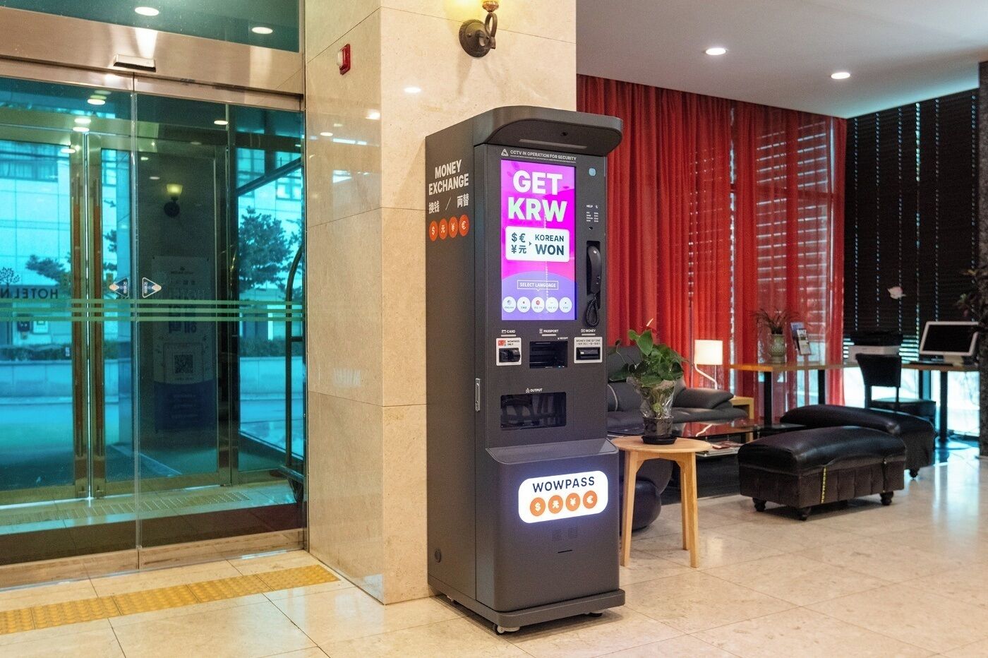Un distributeur de billets permettant d’échanger des devises, agrémenté d’une pancarte indiquant « Obtenez des dollars néo-zélandais », situé dans un hall d’entrée avec un rideau rouge et un espace détente.
