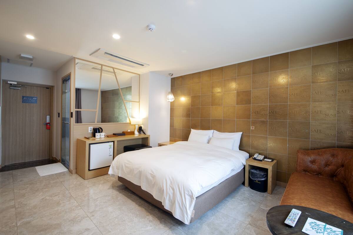 A hotel room with a bed, bedside table, lamp, and a brown leather sofa.