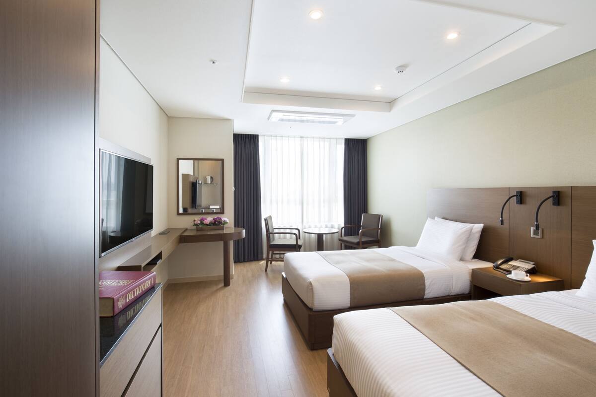 A hotel room with two beds, a desk with a flat-screen TV, a chair, and a small table.