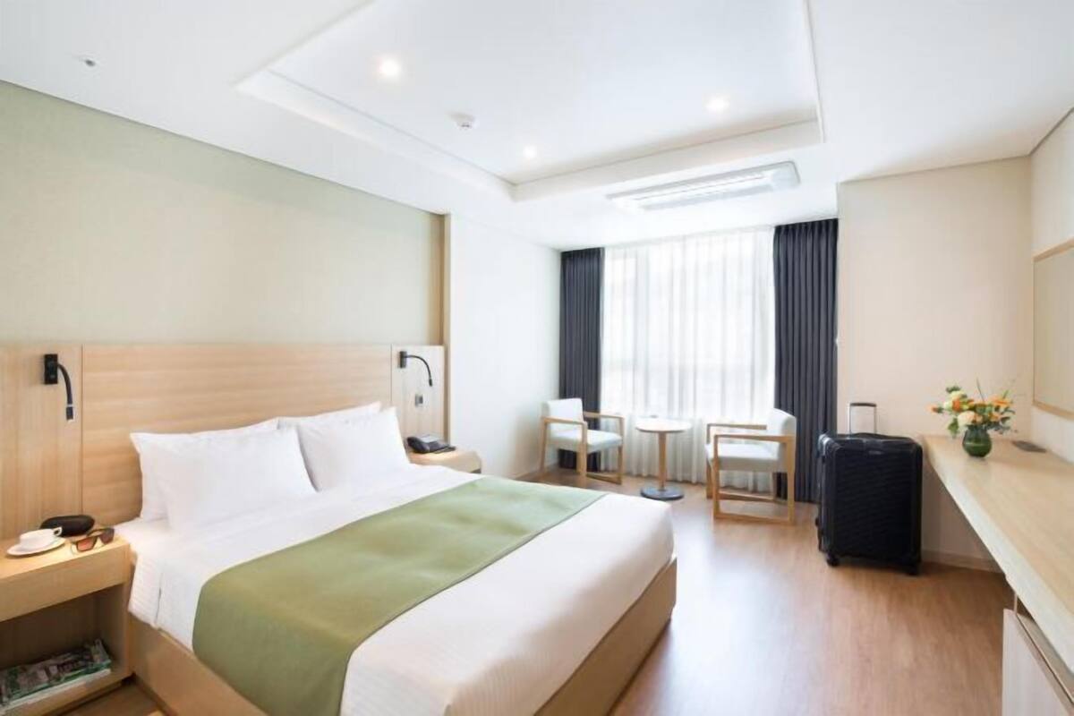 A hotel room with a bed, bedside table, lamp, chair, desk, and a suitcase.