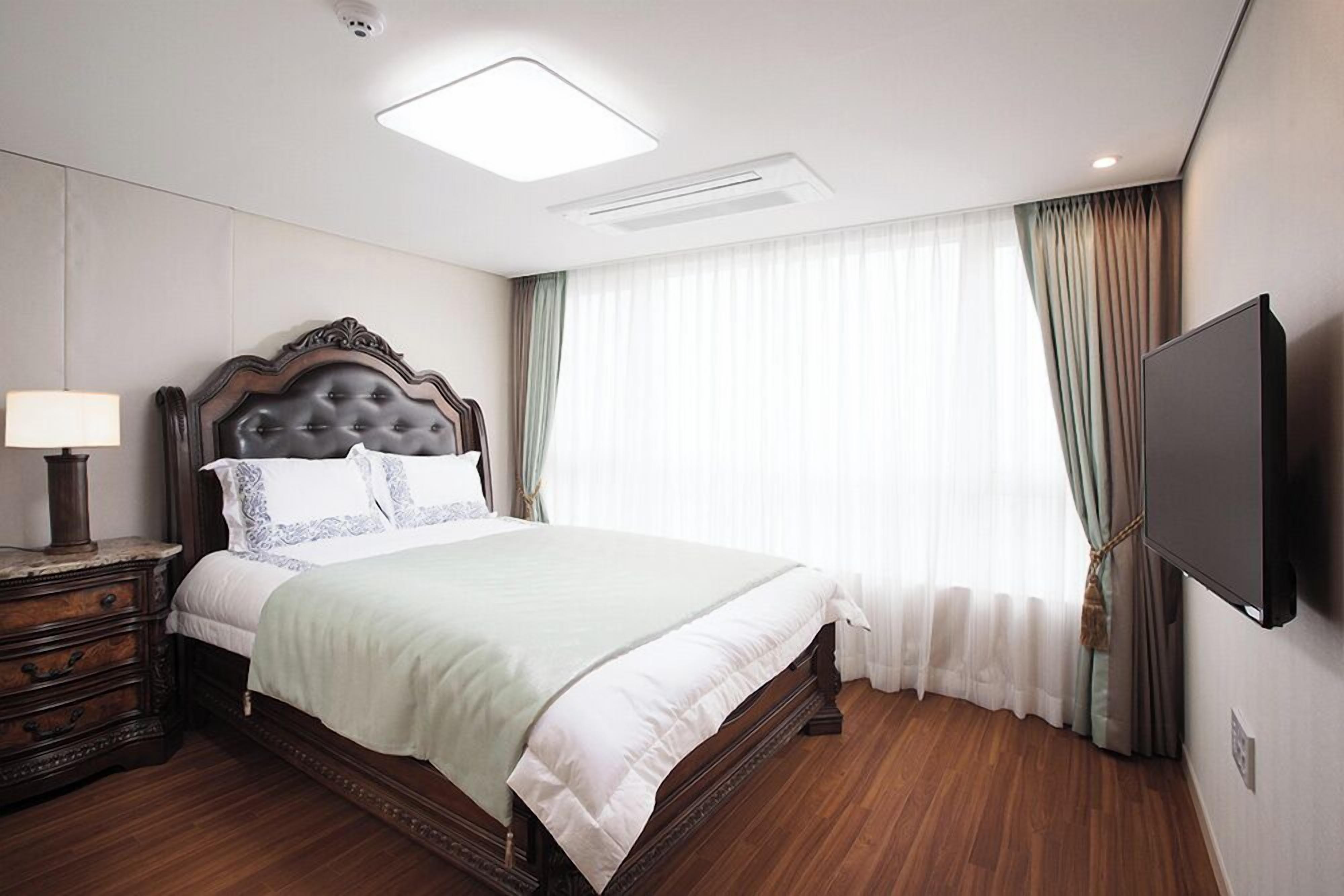 Un dormitorio con una cama grande, una mesita de noche con lámpara, un televisor de pantalla plana en la pared y una ventana con cortinas translúcidas.