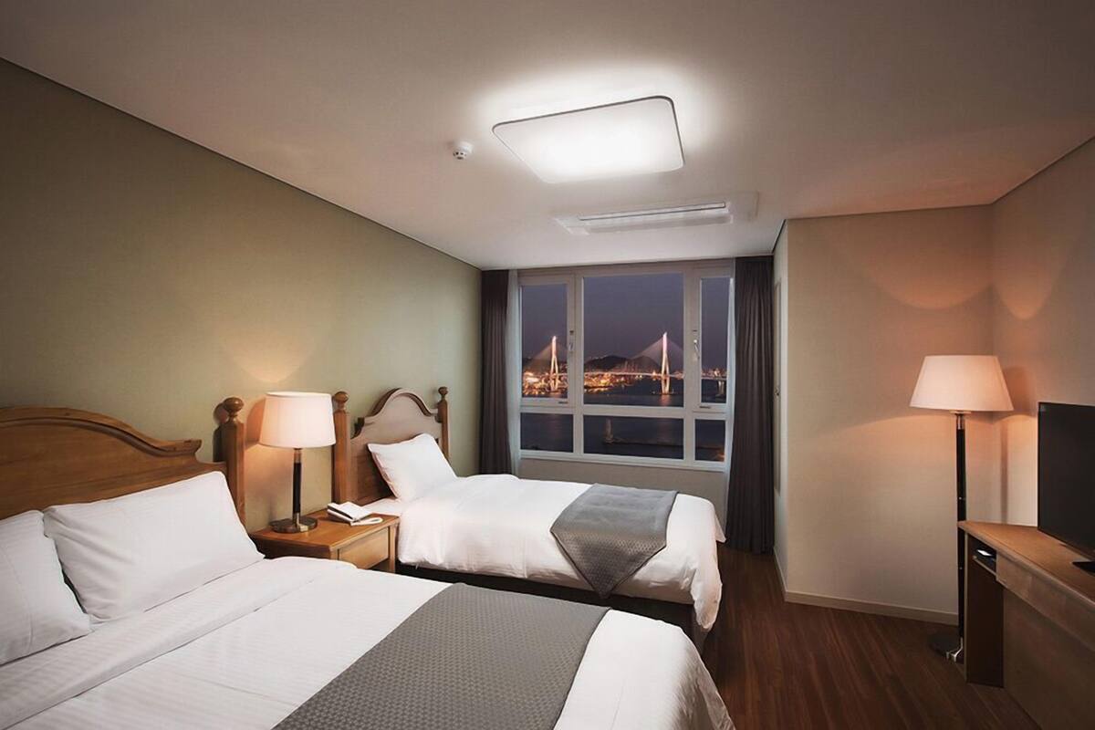 A hotel room with two beds, a nightstand with a lamp, a TV stand, and a window with a city view.