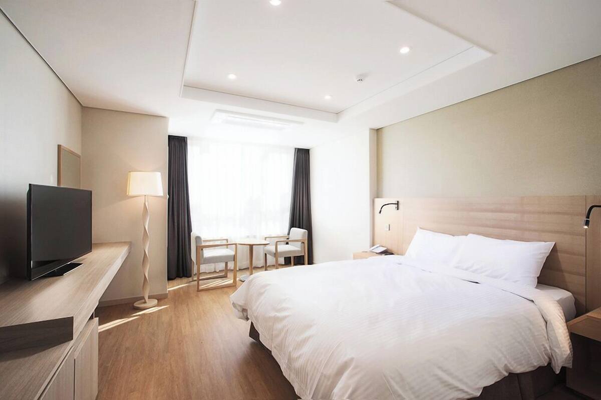 A hotel room with a bed, bedside table, lamp, TV, and a desk with a chair.