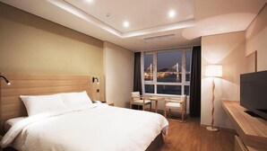 A hotel room with a large bed, a desk with a TV, a chair, a lamp, and a view of a cityscape at night. - Crown Harbor Hotel Busan (Busan)