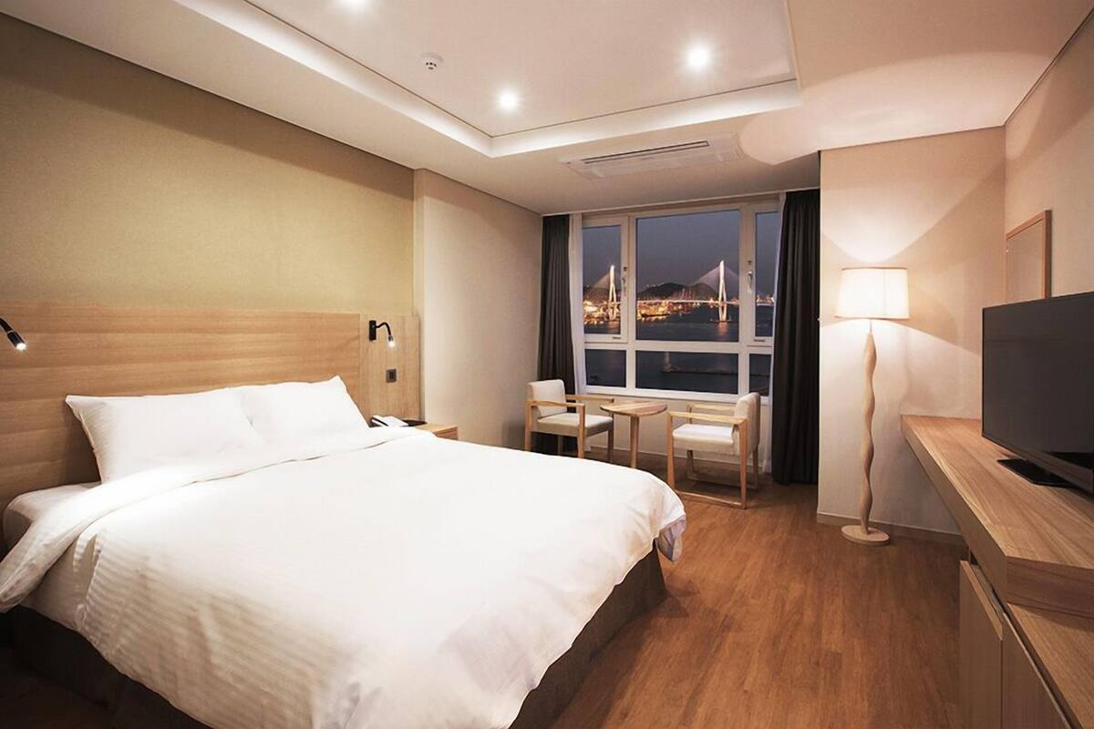 A hotel room with a large bed, a desk with a TV, a chair, a lamp, and a view of a cityscape at night.