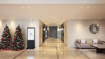 A modern hotel lobby with two decorated Christmas trees, a reception desk, a couch, and a wall-mounted wreath.