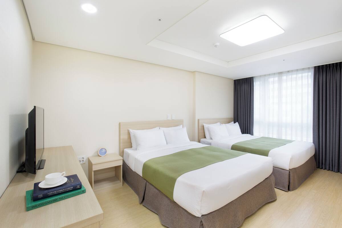 A hotel room with two beds, a desk with a cup and books, a TV, and a window with curtains.