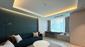A modern living room with a teal accent wall, a dark blue sofa, a round coffee table, and a view of the city through large windows. - Crown Harbor Hotel Busan (Busan)