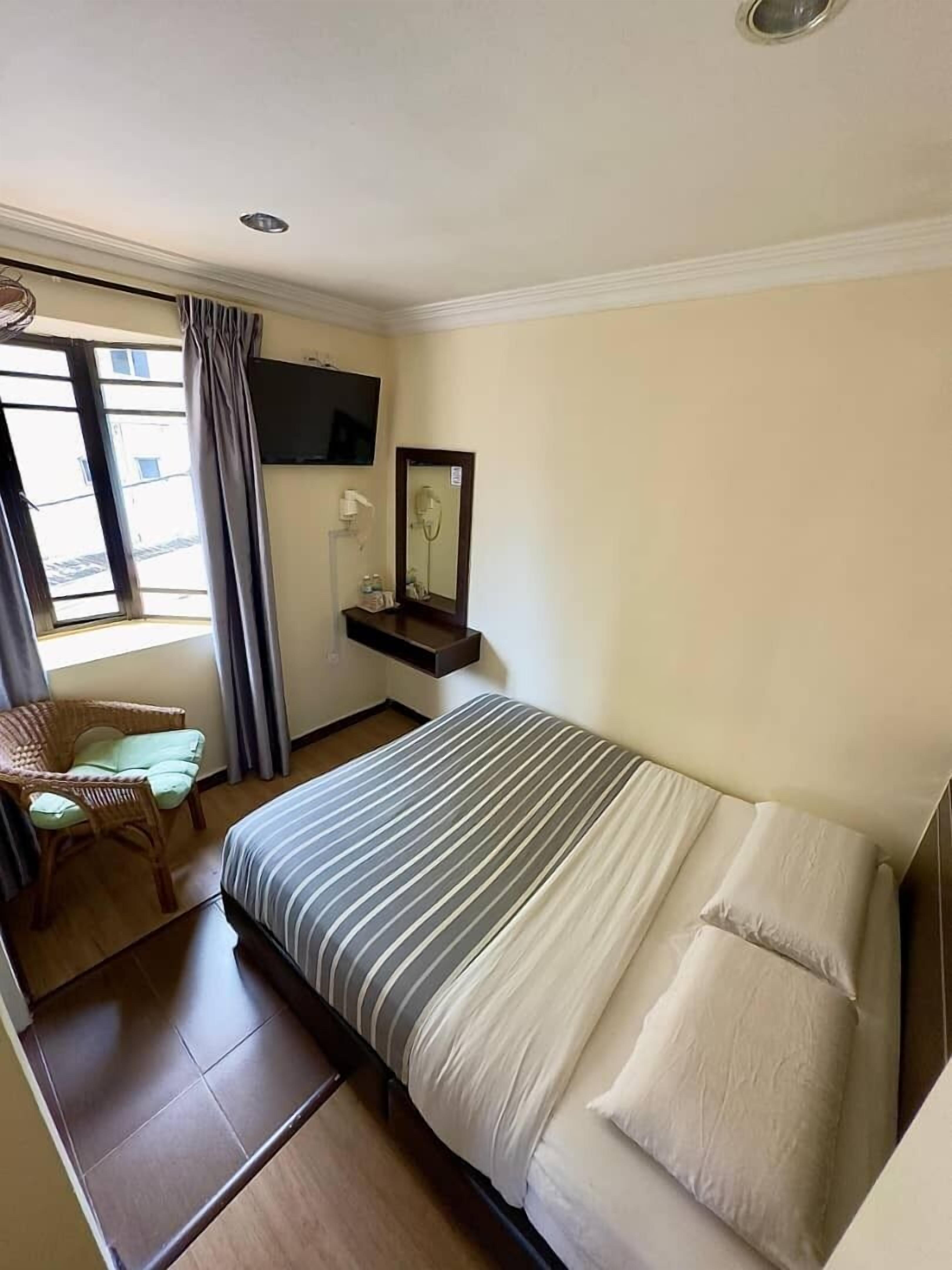 Una habitación de hotel con una cama, una silla, un televisor, un espejo y un ventanal con cortinas.