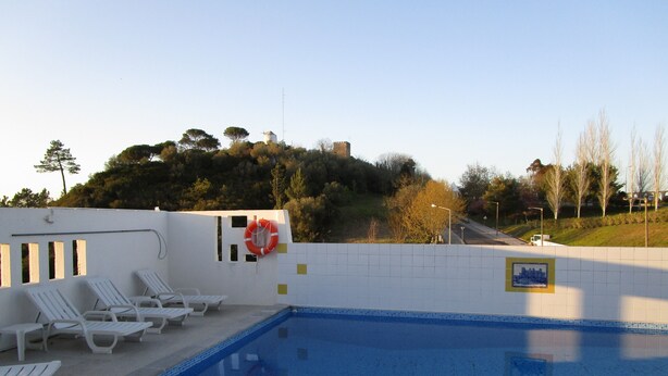 A swimming pool with white lounge chairs, a lifebuoy, and a tiled wall with a cityscape poster. - Hotel Louro (Obidos)