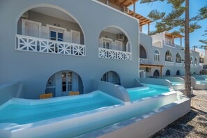 A building with multiple balconies, each with a white railing and a window. There are outdoor swimming pools in front of the building. - Maistros Village (Santorini)