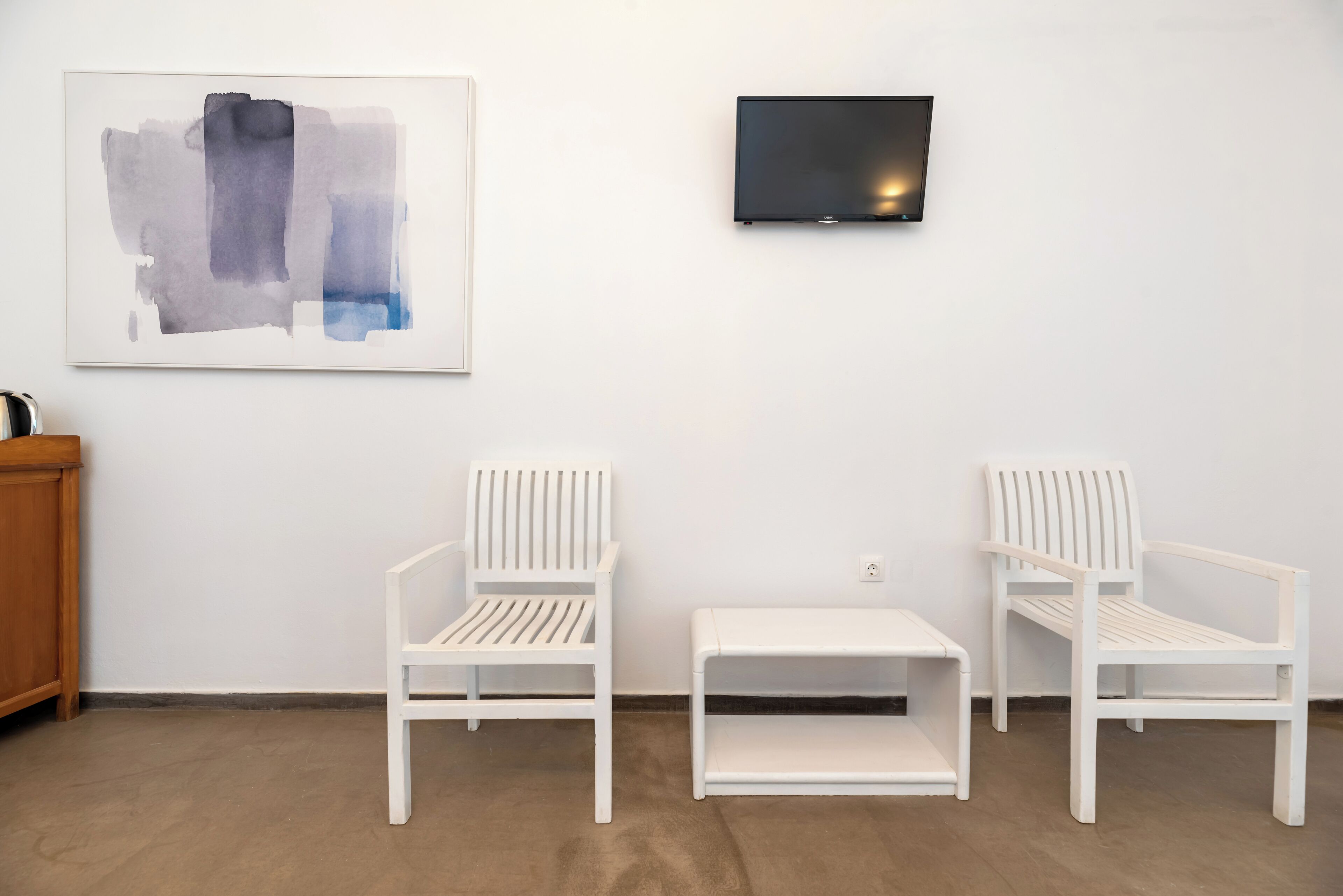 A minimalist room with two white chairs, a small white table, a framed abstract painting, and a mounted flat-screen TV.