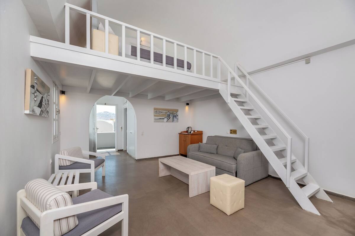 A spacious living area with a high ceiling, a staircase, a grey sofa, a wooden cabinet, and a coffee table.
