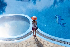A person standing by a pool with a dolphin, a stone tiled deck, and a clear blue sky. - Maistros Village (Santorini)