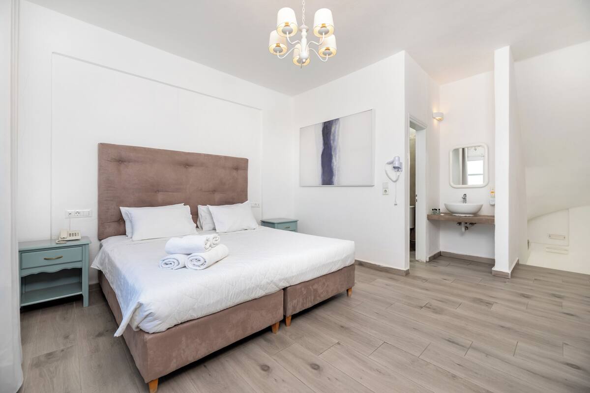 A bedroom with a large bed, bedside tables, a nightstand, a chandelier, a painting, a bathroom with a sink and mirror.