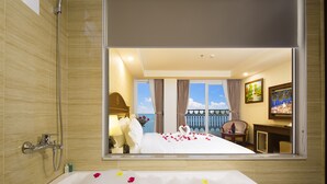Quarto de hotel com uma banheira grande cheia de pétalas de rosa, chuveiro, cama com vista para o mar e varanda com guarda-corpo.