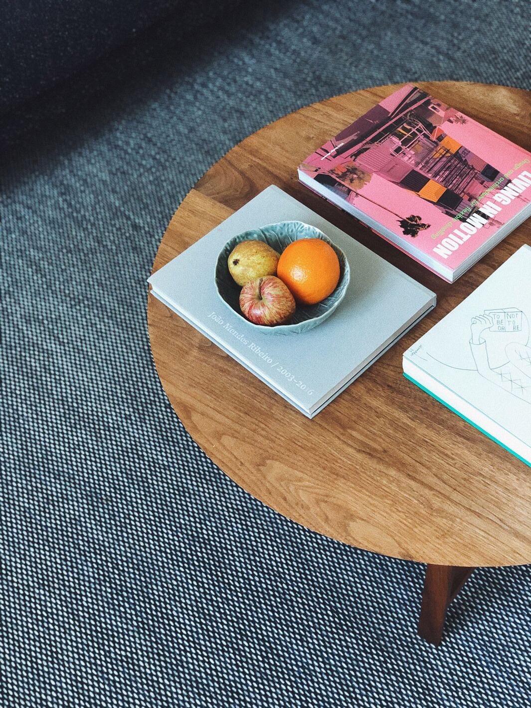 A round wooden table with a bowl containing apples and an orange, a book titled "TO BE OR NOT TO BE OR RE", and a sketchbook.