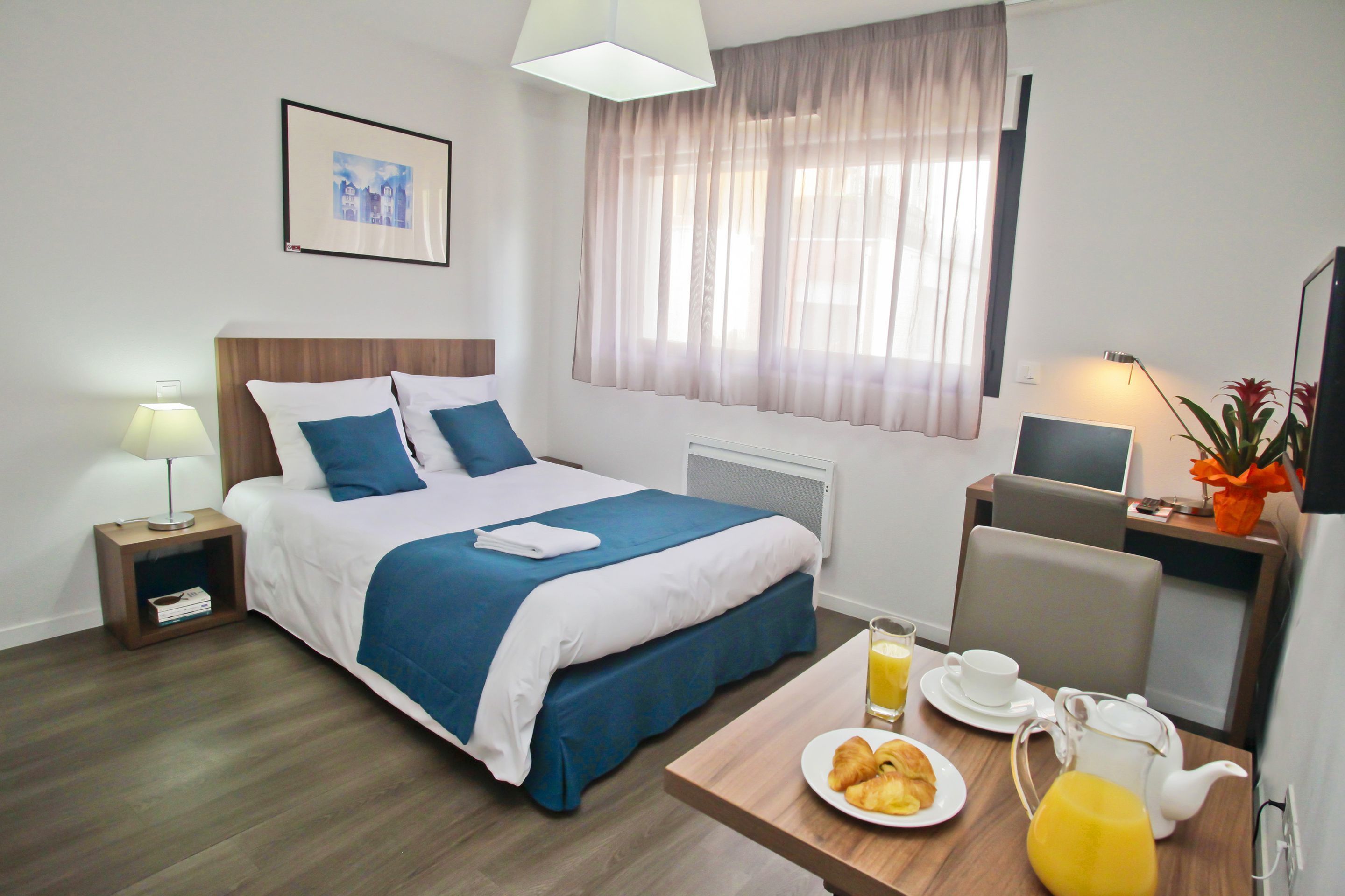A hotel room with a bed, bedside table, lamp, and a breakfast table with croissants and orange juice.