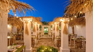 An outdoor dining area with tables and chairs, surrounded by columns and thatched roofs, under string lights. - Masseria Narducci (Fasano)