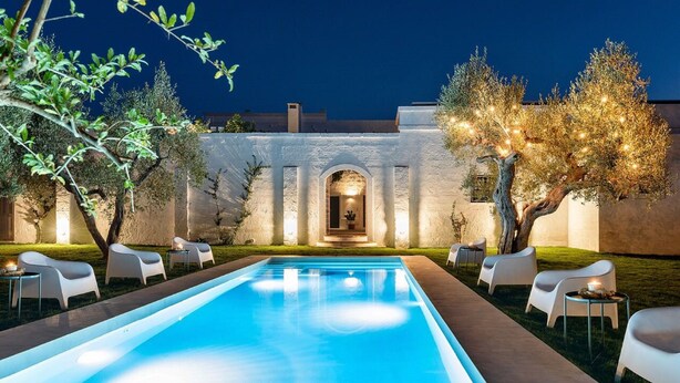 A swimming pool with white lounge chairs and tables, surrounded by olive trees and a white building with arches. - Masseria Narducci (Fasano)