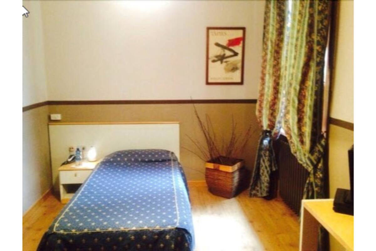 Habitació petita i senzilla amb un llit, una tauleta de nit, una làmpada, un quadre al mur i una finestra amb cortines.