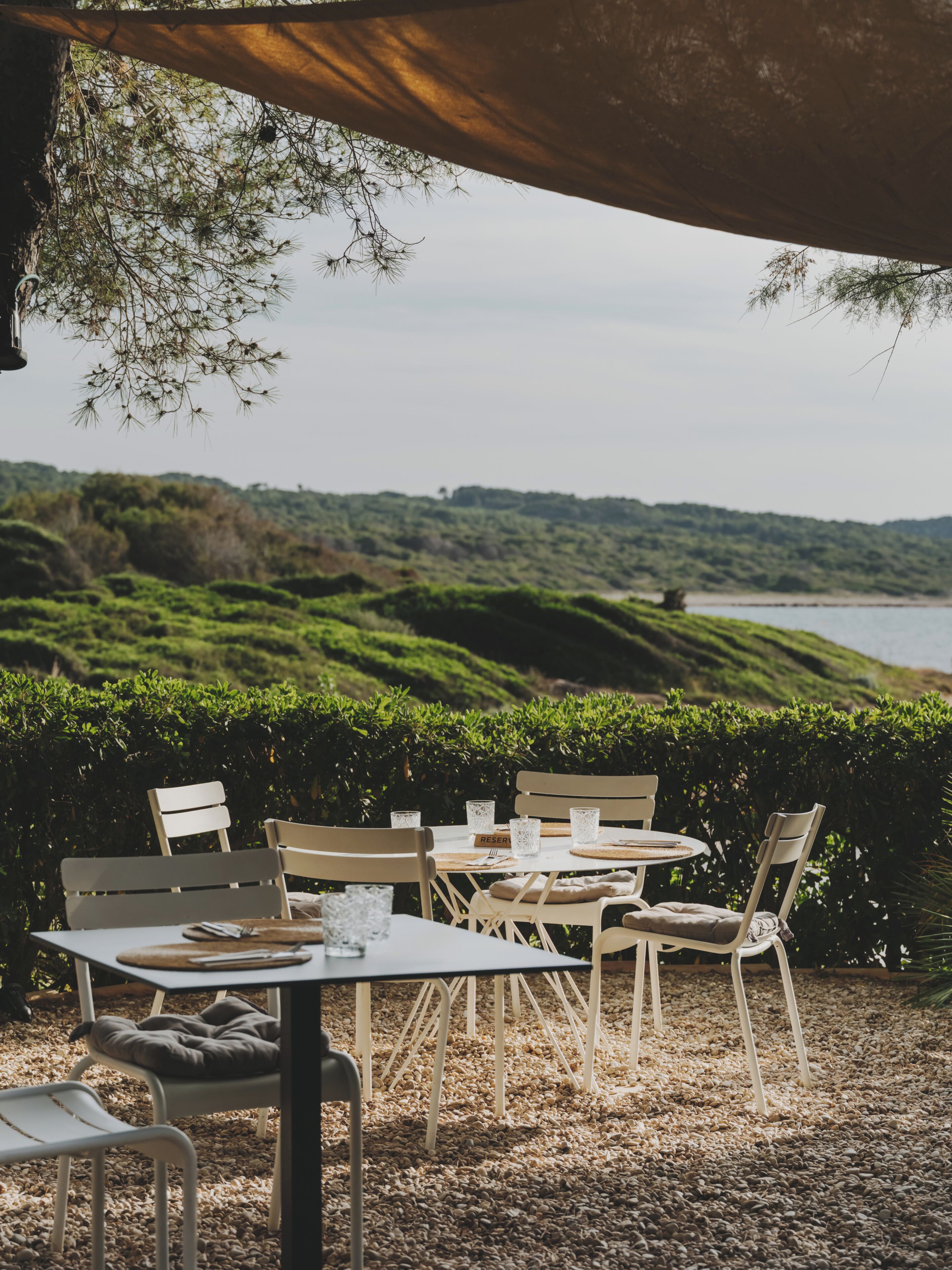 Un área de comedor al aire libre con mesas y sillas, vista a una colina y un cuerpo de agua al fondo.