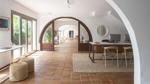 Una habitación amplia con suelo de baldosas, un escritorio con computadora y una silla. Hay otomanas y una alfombra en el suelo.