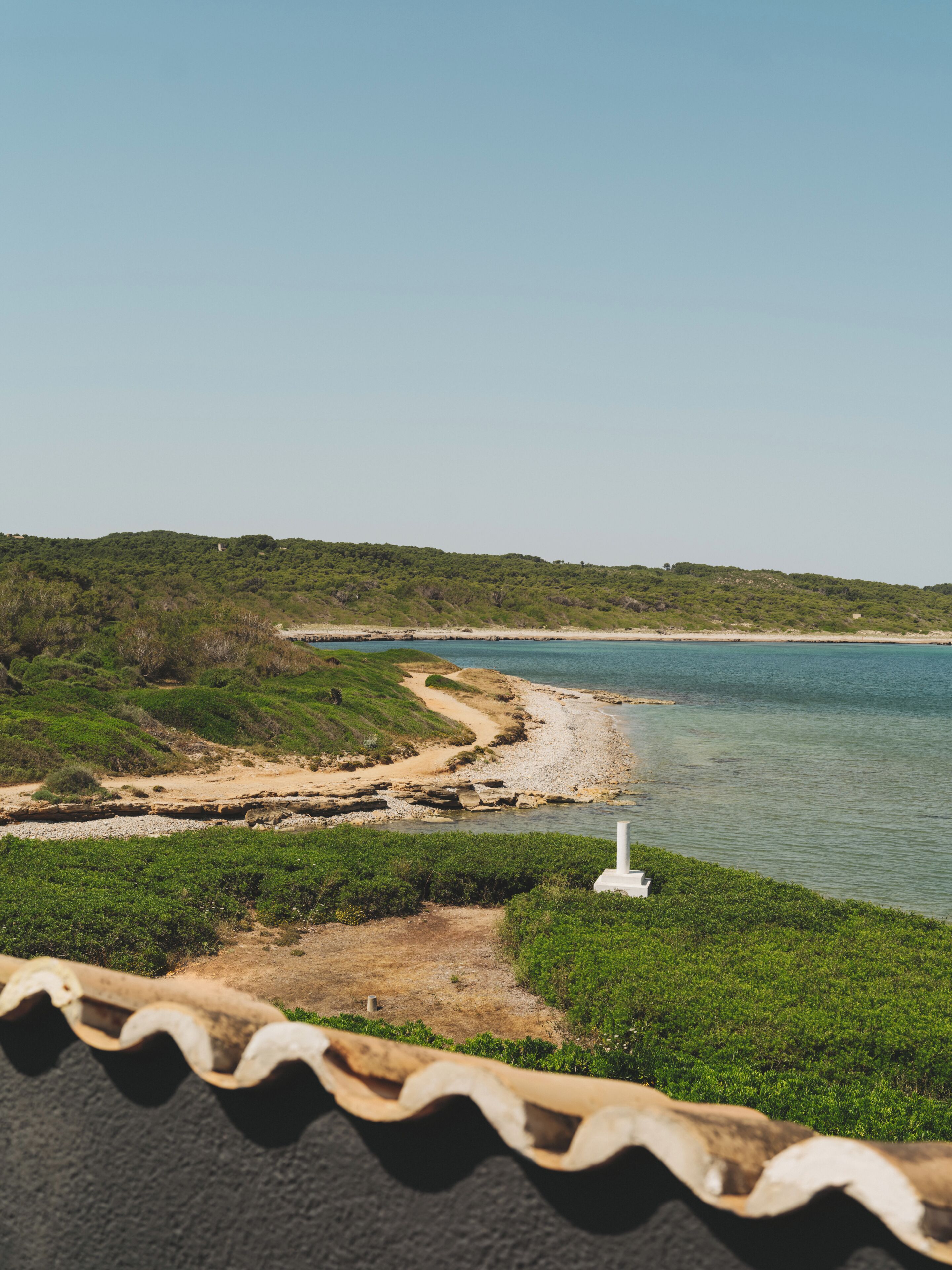A coastal landscape with a white structure, possibly a lighthouse, near a sandy beach with green vegetation.