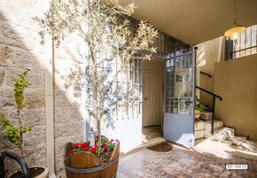 Un patio con un muro de piedra, una jardinera de madera con flores rojas, un árbol alto y una escalera que lleva a un nivel superior.