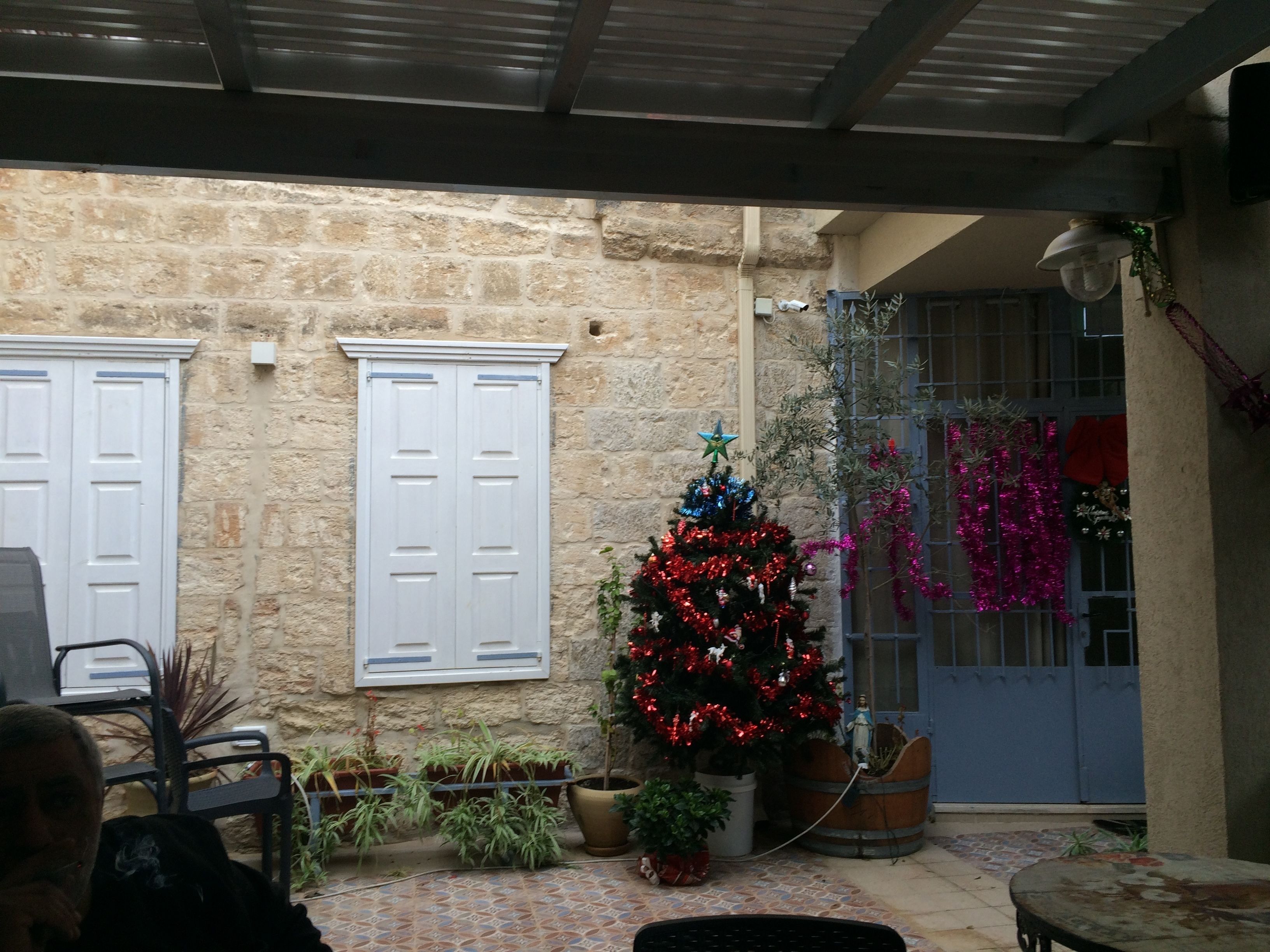 Un bâtiment en pierre avec des volets blancs, un sapin de Noël décoré, des plantes en pot et une porte bleue.